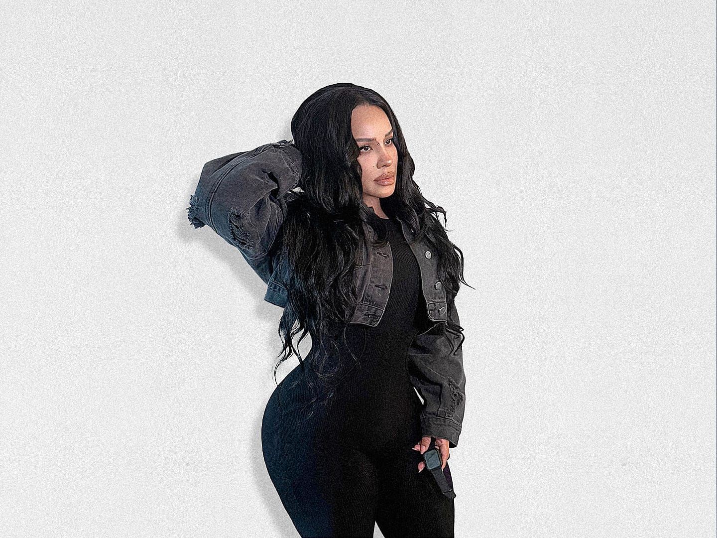 Full-body fashion portrait of a woman with long, wavy black hair wearing a fitted black outfit layered with a cropped charcoal denim jacket. She poses against a minimalist white wall with one hand in her hair, showcasing an understated monochrome look inspired by elevated everyday style.