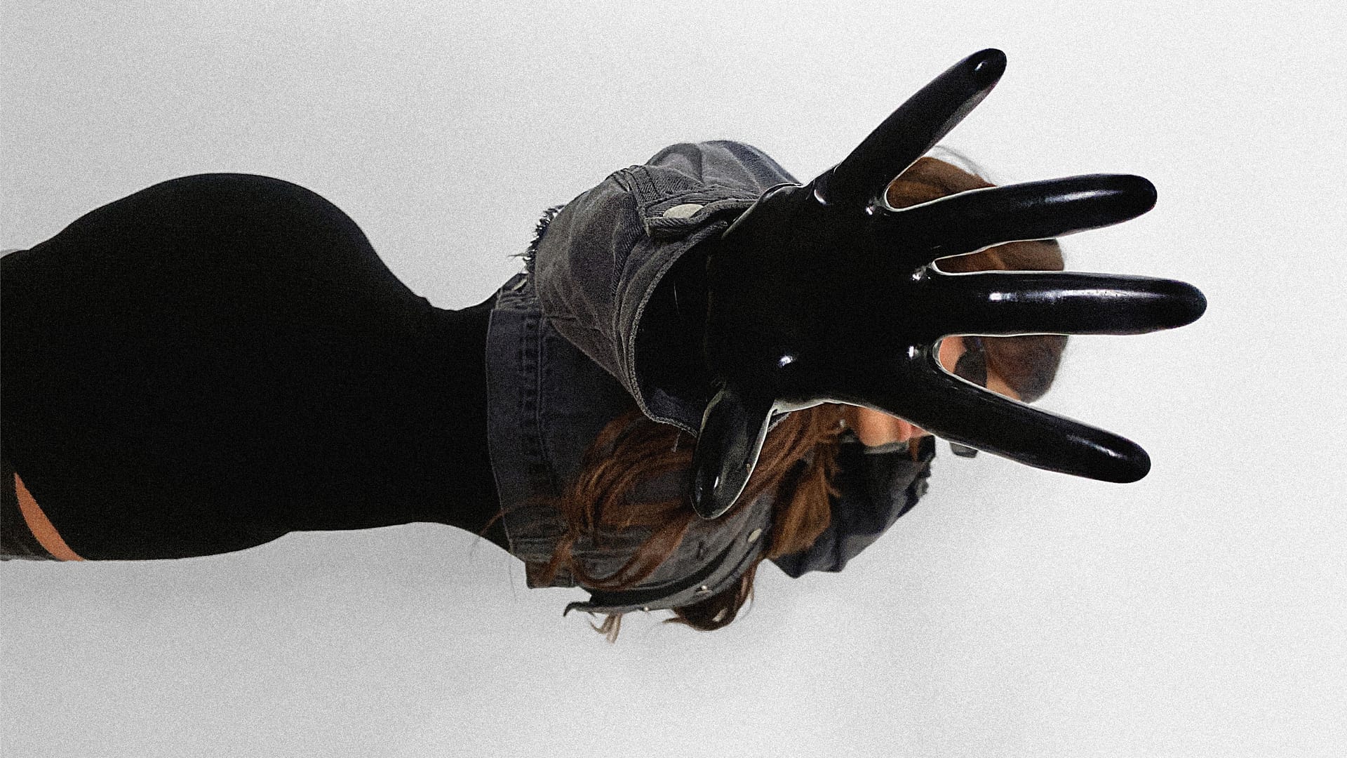 Editorial fashion image of a woman wearing black latex gloves and a dark jacket, extending her hand toward the camera in a dramatic, high-contrast pose.