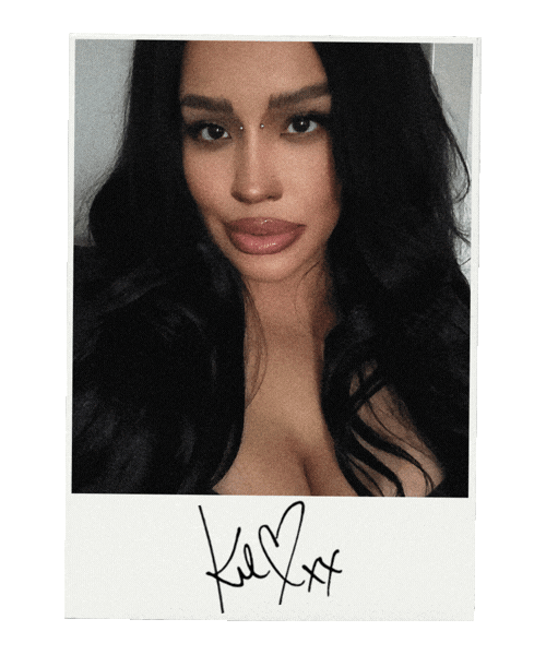**Alt text:**
Polaroid-style portrait of a woman with long black wavy hair, glossy lips, and bridge piercings, posing against a neutral background. The photo features a handwritten signature on the white border beneath the image.