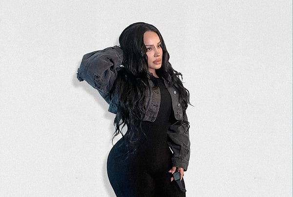 Full-body fashion portrait of a woman with long, wavy black hair wearing a fitted black outfit layered with a cropped charcoal denim jacket. She poses against a minimalist white wall with one hand in her hair, showcasing an understated monochrome look inspired by elevated everyday style.