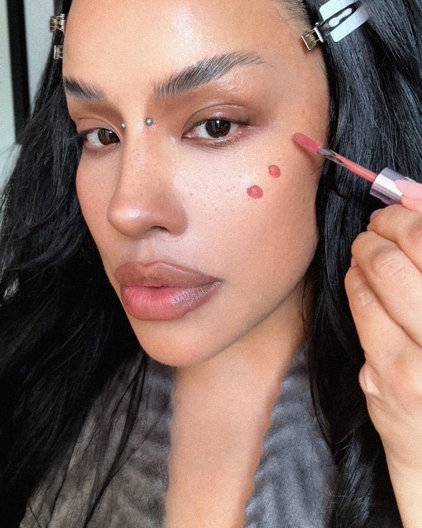 Close-up of a woman with long black hair and bridge piercings applying pink liquid blush to her cheek with a makeup applicator, wearing hair clips and minimal makeup for a beauty tutorial-style moment.